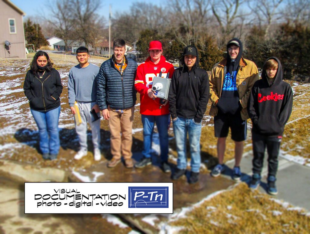 Instructor Scott Hughes poses with high school students students, one of which is holding a camera.