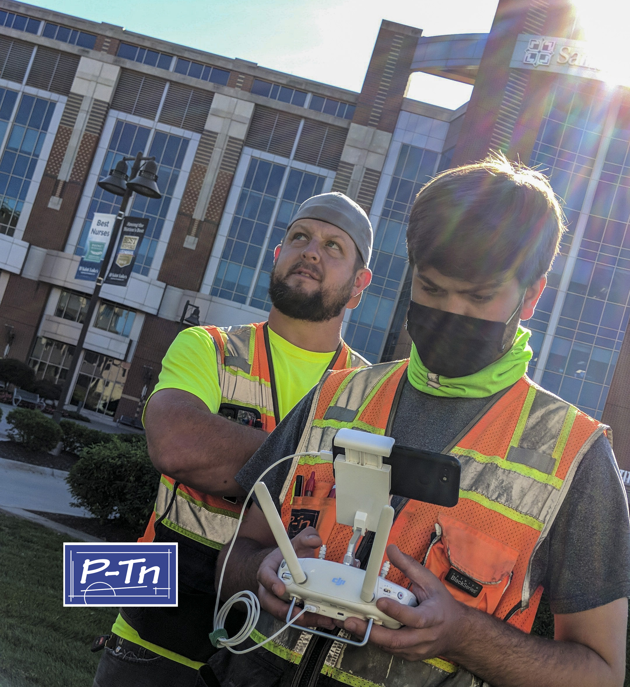 Two men in bright orange vests using a drone control. | P-Tn