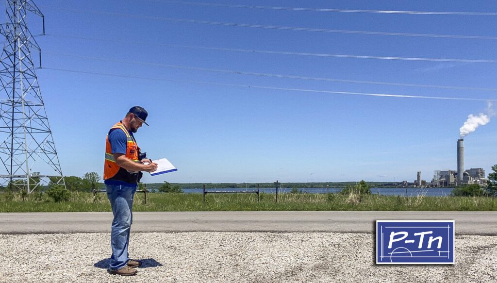 man in construction vest stands on road writing on paper with power plant in background | P-Tn