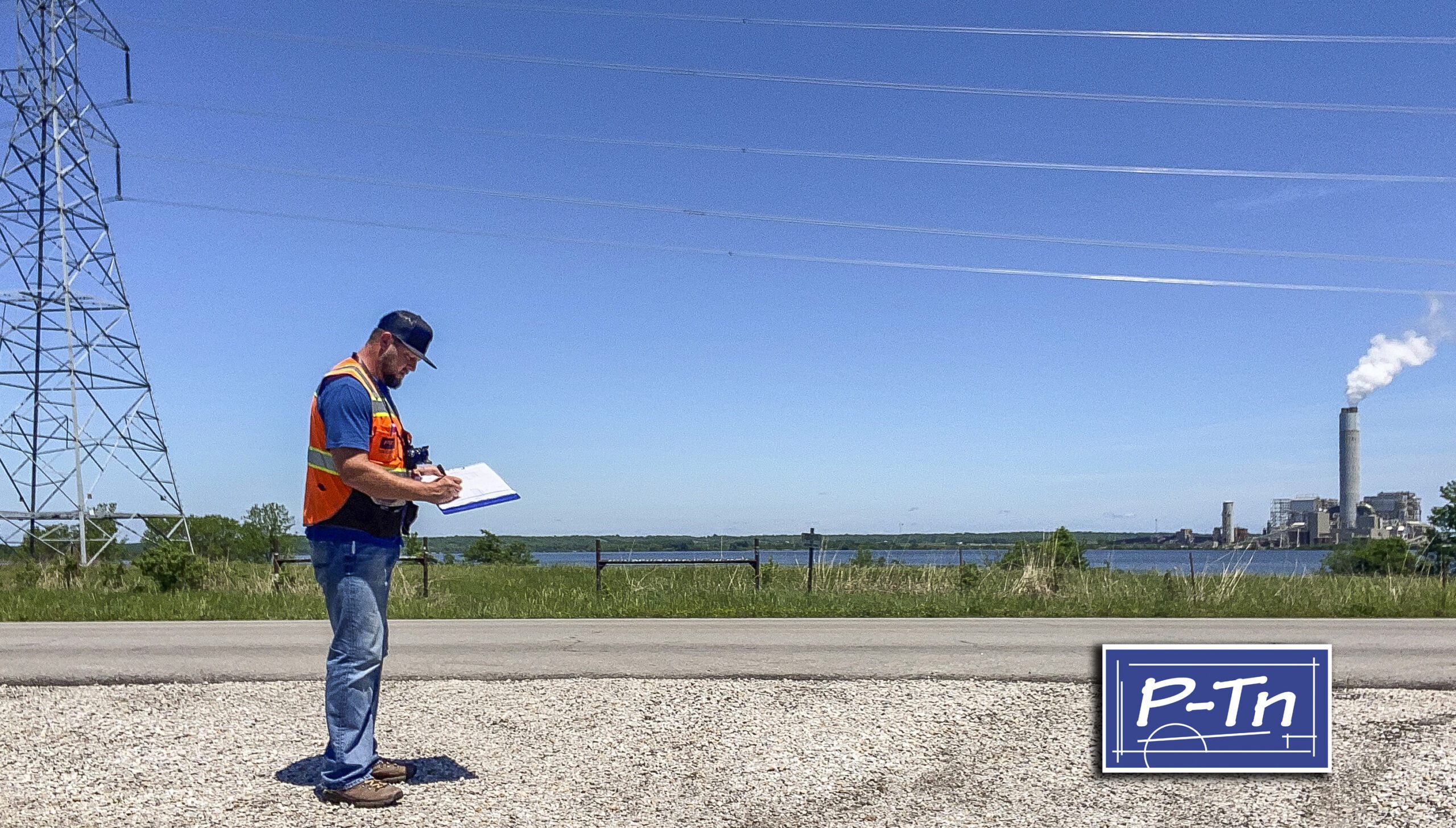 man in construction vest stands on road writing on paper with power plant in background | P-Tn