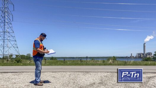 man in construction vest stands on road writing on paper with power plant in background | P-Tn